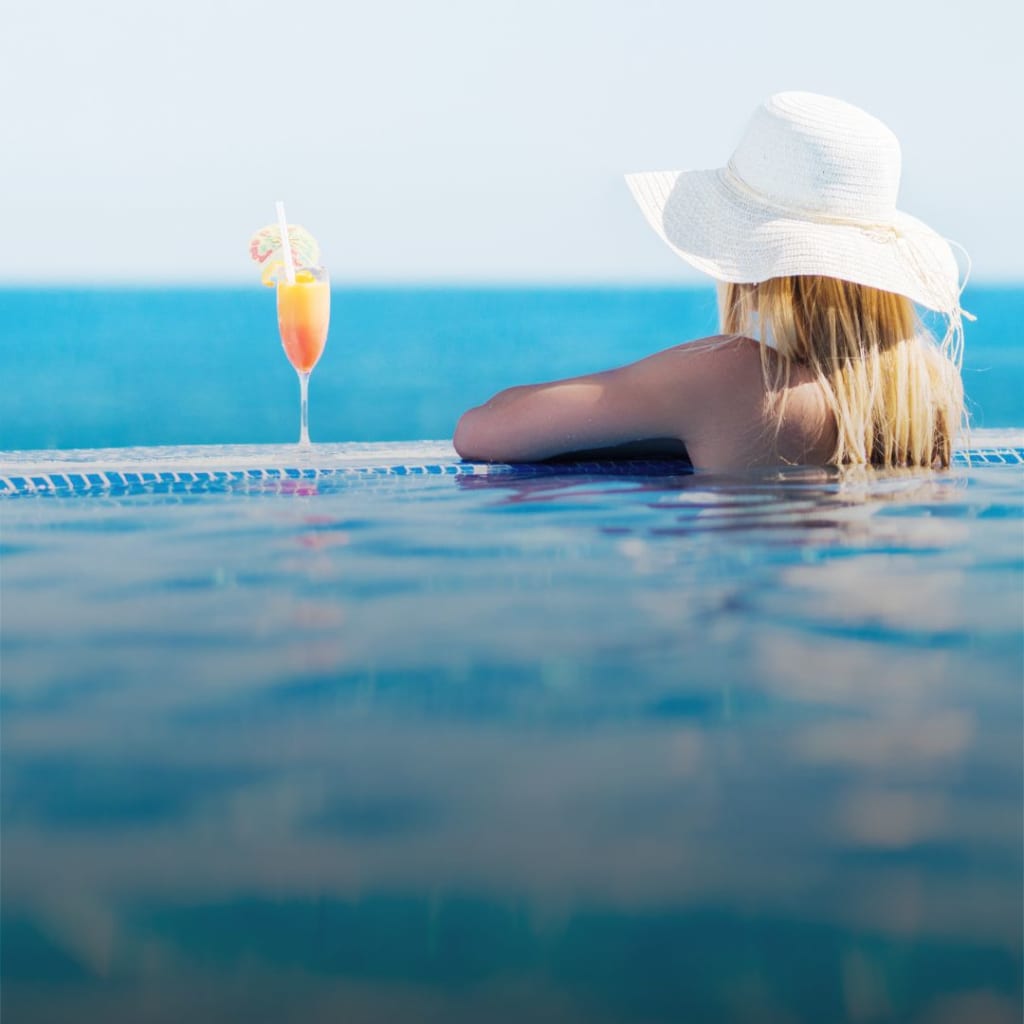 Rear view of a woman enjoying in summer day at the swimming pool and looking at view. Summer cocktail is next to her.